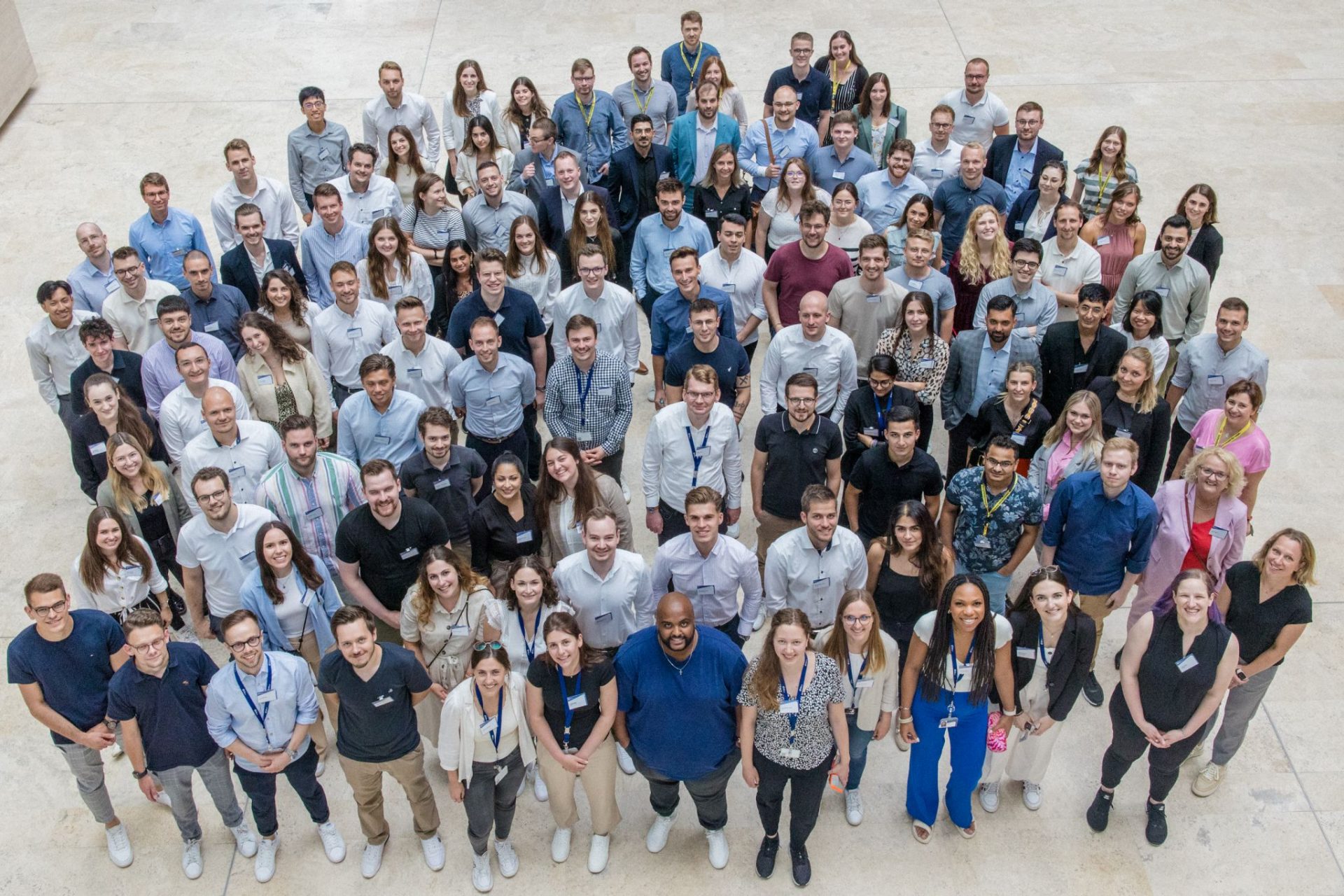 Das Bild zeigt eine große Gruppe von Menschen, die zusammenstehen und in die Kamera lächeln. Es könnte eine Firmenveranstaltung, ein Workshop, eine Konferenz oder ein Teammeeting sein, bei dem Gruppenfotos gemacht werden, um die Teilnehmer zu dokumentieren oder für Erinnerungszwecke festzuhalten. Die Vielfalt der Kleidung und der entspannte Ausdruck auf den Gesichtern deuten darauf hin, dass es sich um eine informelle oder halbformale Veranstaltung handelt.