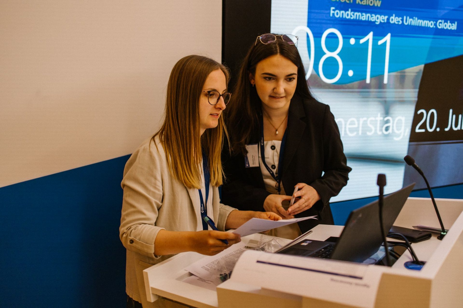 Das Foto zeigt eine Umgebung in einem Büro oder Konferenzraum mit technischer Ausstattung, einem Tisch mit einem Laptop und Mikrofonen darauf. Eine Präsentation oder ein digitales Display ist im Hintergrund sichtbar, auf der Informationen über eine Veranstaltung angezeigt werden, darunter Datum und Uhrzeit.
