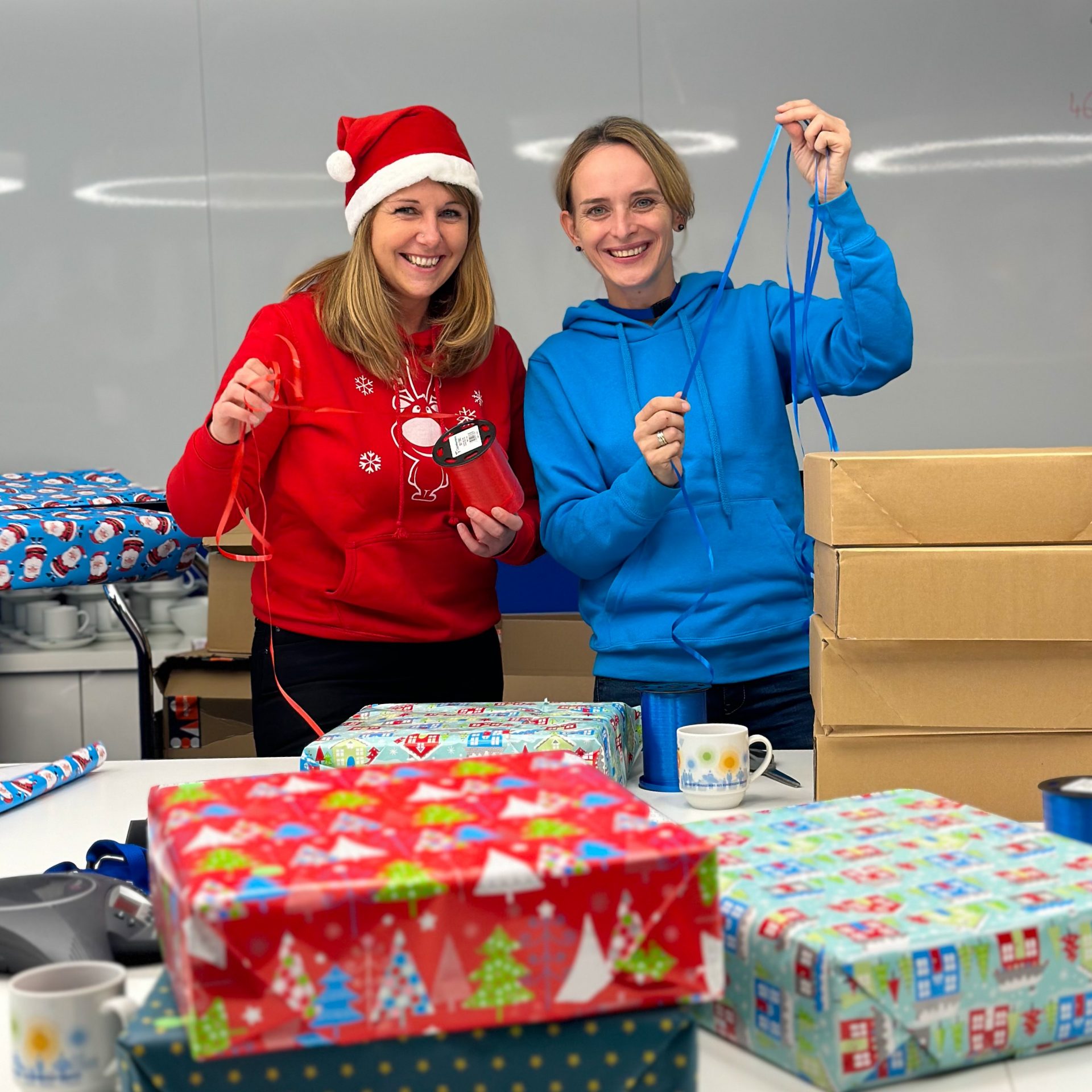 Das Bild zeigt eine Umgebung, die auf das Verpacken von Geschenken während der Weihnachtszeit hinweist. Es gibt eine Sammlung von farbig eingewickelten Geschenken, Geschenkpapierrollen und Tassen auf einem Tisch. Im Hintergrund sind größere Kartons sichtbar.