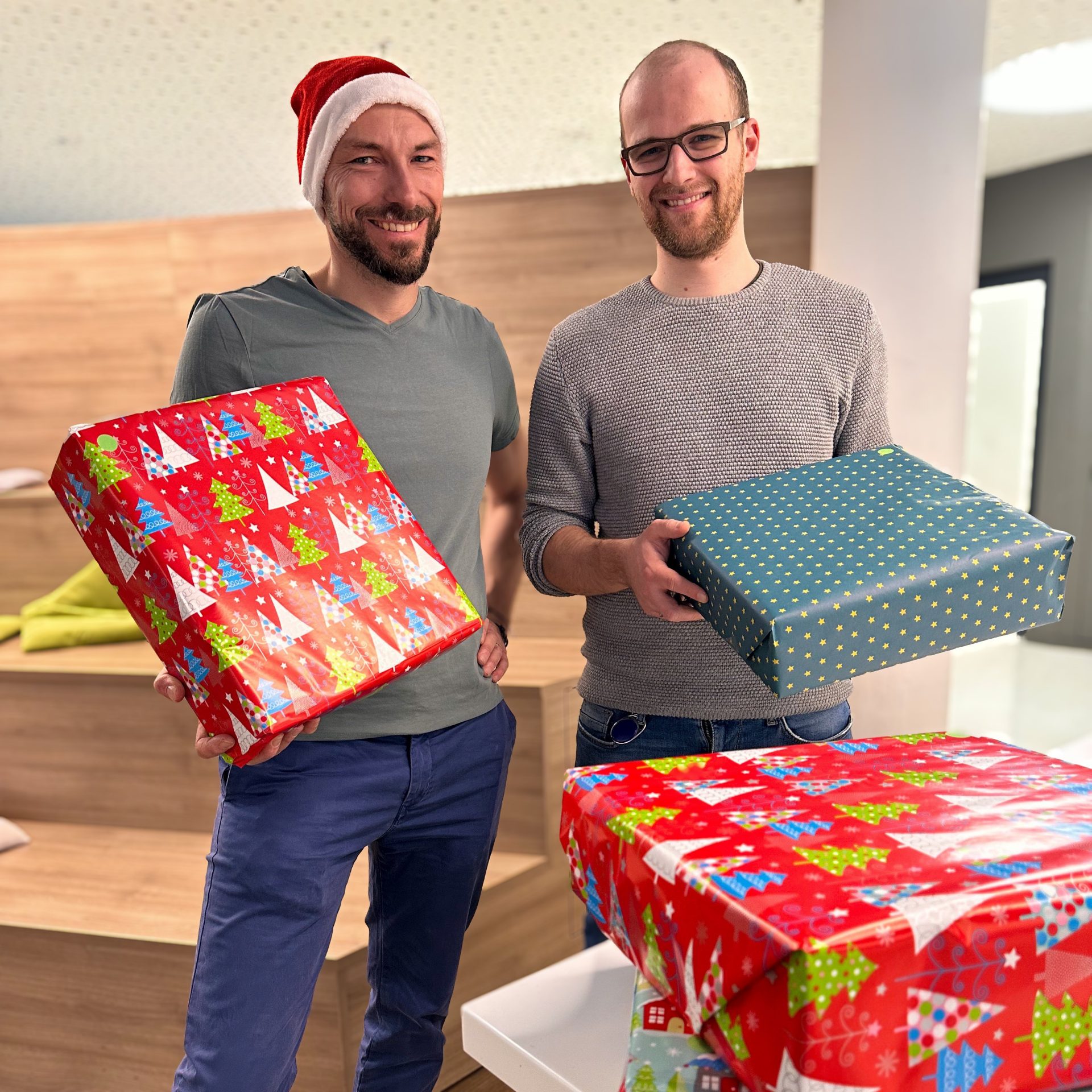 Das Bild zeigt zwei Personen, die Geschenkboxen halten, möglicherweise im Rahmen einer Weihnachtsszene, da einer eine Weihnachtsmütze trägt und die Boxen weihnachtlich verpackt sind.