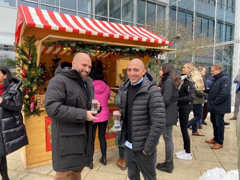 Das Bild zeigt eine Gruppe von Menschen, die draußen vor einem kleinen Weihnachtsmarktstand stehen. Der Stand ist mit Weihnachtsdekorationen geschmückt und hat ein rot-weiß gestreiftes Dach. Einige der Menschen halten Gläser oder Tassen in ihren Händen.