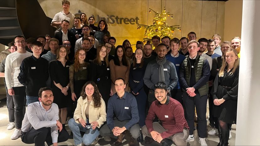 Das Bild zeigt eine Gruppe von Menschen, die zusammen für ein Gruppenfoto posieren. Die Gruppe steht vor einem Hintergrund mit einem wandmontierten Logo mit der Aufschrift "50Street" und einem gelb beleuchteten Baum. Einige Teilnehmer tragen Namensschilder auf ihrer Kleidung, was auf eine organisierten Veranstaltung oder ein Treffen hindeutet.