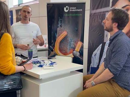 Das Bild zeigt eine Umgebung, die aussieht wie eine Messe oder eine Veranstaltung mit einem Infostand. Auf dem Stand ist eine Werbetafel von "Union Investment" zu sehen, die ein abgebildetes rotglühendes Hufeisen zeigt. Es könnten Informationsmaterialien und Schreibutensilien auf dem Tisch sein.
