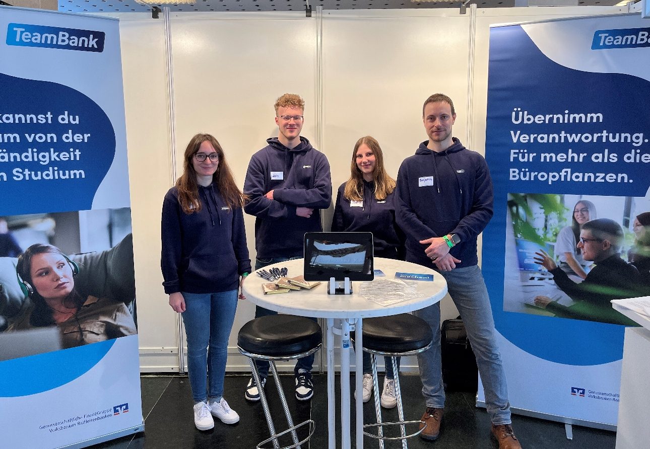 Das Bild zeigt eine Gruppe von vier Personen, die vor zwei TeamBank-Bannern stehen. Diese wirken wie Teilnehmer einer Messe oder einer Veranstaltung. Neben ihnen steht ein Tisch mit einem Laptop darauf. Die Banner der TeamBank enthalten Text, der zu mehr Eigenständigkeit im Studium und zur Übernahme von Verantwortung auffordert. "Übernimm Verantwortung. Für mehr als die Büropflanzen."
