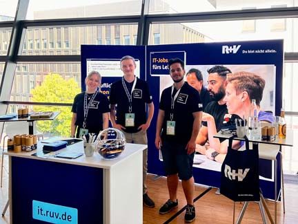 Das Bild zeigt einen Messestand von R+V, auf dem Informationstafeln über IT-Jobs und Lernmöglichkeiten präsentiert werden. Drei Personen stehen an diesem Stand, der mit Broschüren, einem Laptop und anderen Präsentationsmaterialien ausgestattet ist. Im Hintergrund sind zwei beidseitige Poster mit weiterführenden Informationen zu sehen.