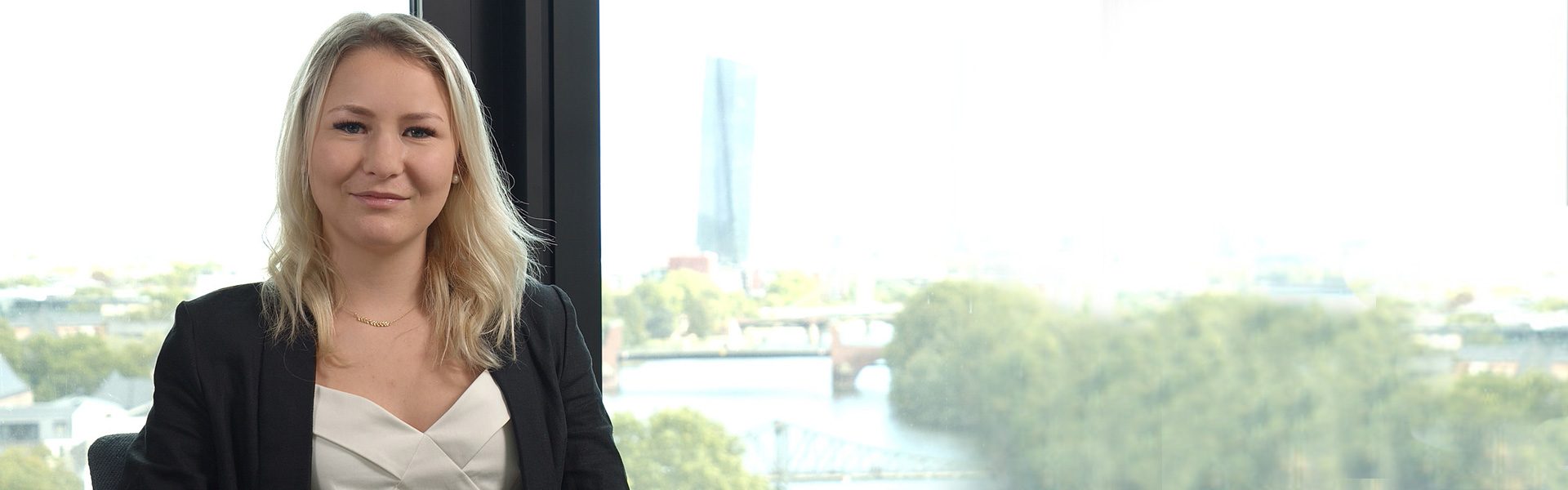 Die Aufnahme zeigt eine Person in formeller Kleidung in einem Büro mit einem großen Fenster, das einen Blick auf eine Stadtlandschaft einschließlich eines Flusses oder einer Wasserfläche bietet.