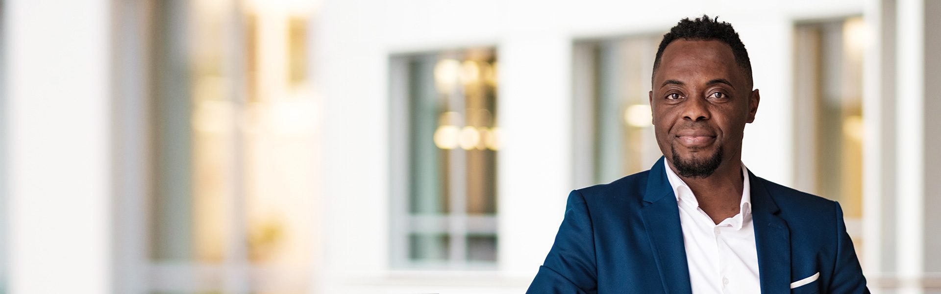 Die Aufnahme zeigt eine Person in einem hellen Bürogebäude mit großen Fenstern und einem eleganten blauen Anzug, einschließlich eines weißen Hemdes darunter, was auf ein professionelles Umfeld hinweist.