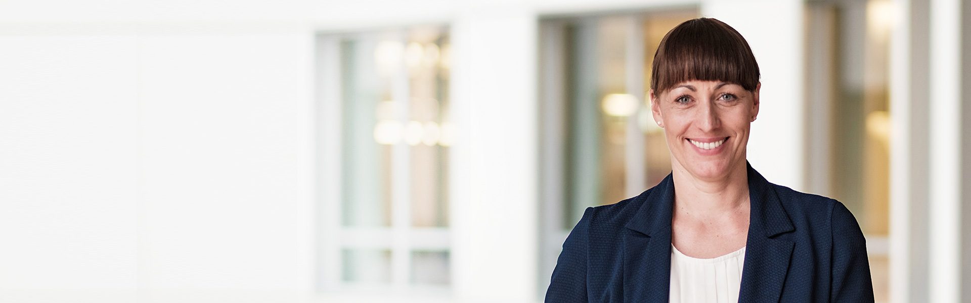 Die Aufnahme zeigt eine Person in formeller Kleidung in einem professionellen oder geschäftlichen Umfeld. Der Hintergrund scheint ein helles Büro oder ein Geschäftsgebäude zu sein, gekennzeichnet durch große Fenster und eine moderne Architektur.