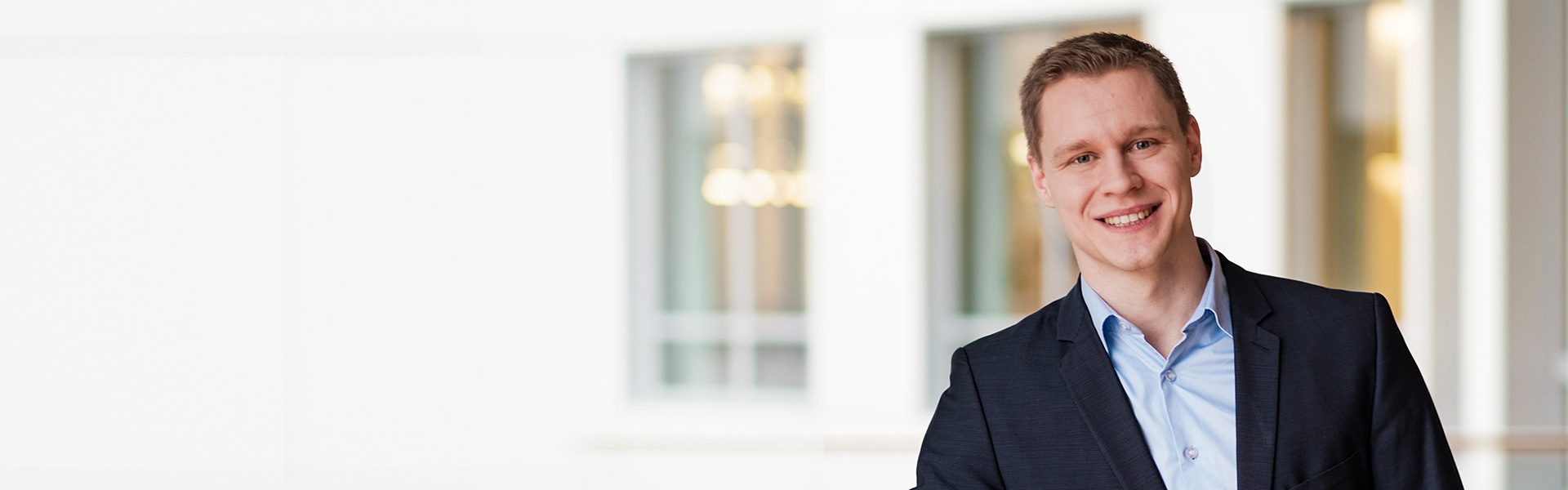 Das Bild zeigt eine Person in einem hellen Büroraum mit großen Fenstern und einem eleganten Holztrimm. Die Person trägt ein formelles Outfit, bestehend aus einem dunkelblauen Anzug und einem hellblauen Hemd.