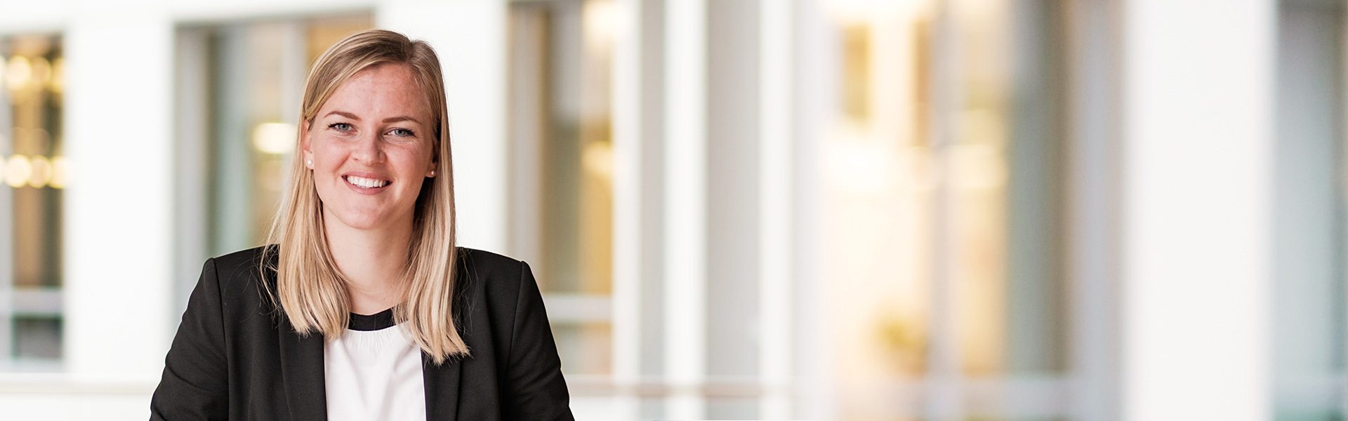 Die Aufnahme zeigt eine Person in formeller Kleidung in einem professionellen oder büroähnlichen Umfeld. Der Hintergrund besteht aus modernen Glas- und Strukturelementen, die auf ein Arbeits- oder Geschäftsumfeld hindeuten.